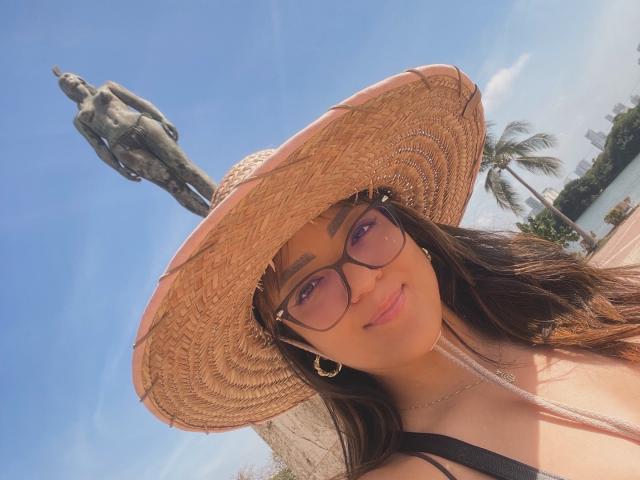 DulceLiz macht ein sonniges Selfie und trägt einen breitkrempigen Strohhut, eine schwarze Brille und Reifenohrringe, im Hintergrund ist eine Statue zu sehen. Hintergrund und klarer blauer Himmel. Ihr dunkles Haar fällt über ihre Schultern..
