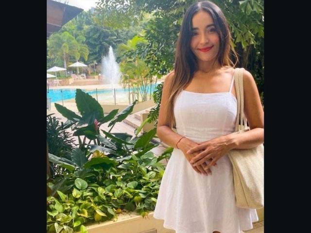 MindyLove stands in a sunny outdoor setting, wearing a white, sleeveless dress with a flared skirt. She has long brown hair, a red lip, and a beige bag. Blue pool in background.