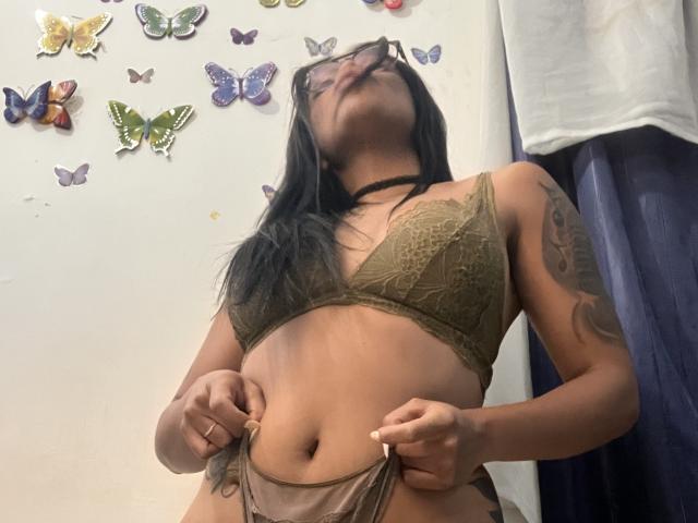 StellaBeckker, a Latina woman with long black hair, wearing a green lace bralette and brown panties, stands with hands on hips, looking up. Butterfly decals on white wall. Tattoo on right arm.