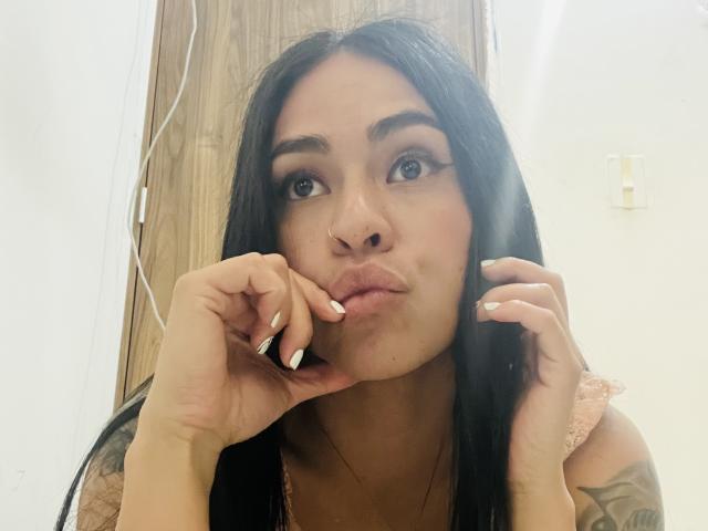 Close-up of StellaBeckker, a Latina woman with long black hair, pouting her lips, touching her face with manicured nails, sporting a nose ring and tattoo on her right arm. Fucking sexy.
