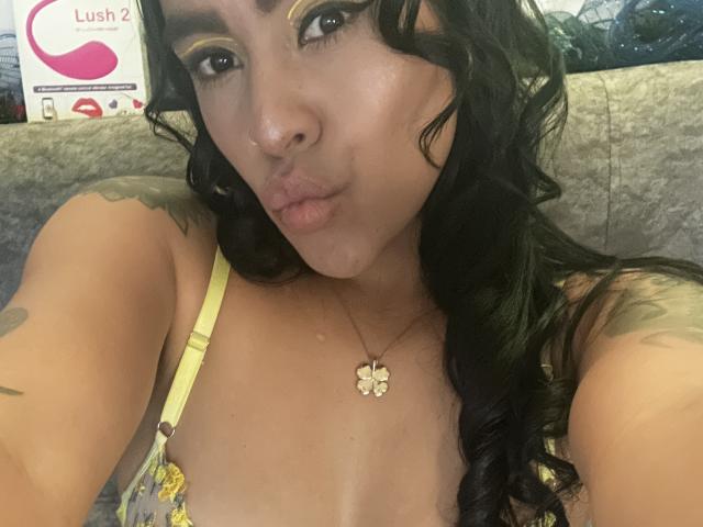 Close-up photo of StellaBeckker, with wet skin, black curly hair, yellow floral top, butterfly necklace, and tattoos on shoulders. StellaBeckker has a seductive look, slightly parted lips, and visible cleavage.
