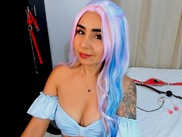 Photograph of EmilyValk with long, pastel blue and pink hair, off-shoulder white top, large cleavage, tattooed arm, smiling, red lips, white bed with red and black straps in background.
