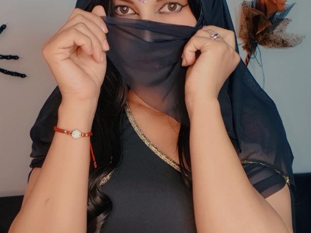 SammyThomsom zieht eine schwarze Maske auf, wodurch braune Augen sichtbar werden. Sie trägt ein schwarzes Oberteil mit goldener Verzierung, ein rotes Armband und einen Ring. Ihre Hände umklammern die Maske verführerisch und erzeugen sexuelle Spannung..