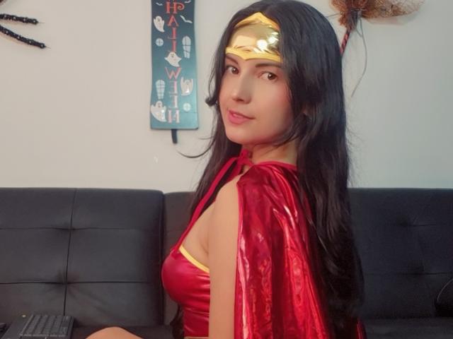 SammyThomsom está sentada em um sofá de couro preto, vestindo uma fantasia vermelha da Mulher Maravilha, com uma tiara dourada e um capa, olhando para... em seu ombro, com uma tensão sexual evidente e um olhar sedutor..