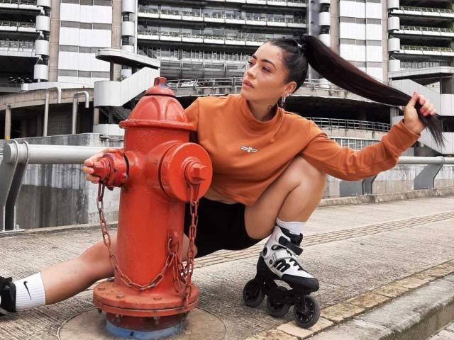 TrinityPrestige, atletisch postuur, hurkt naast een felrode brandkraan, oranje sweatshirt, zwarte korte broek, wit sokken, zwart-witte sneakers, hoge paardenstaart, stedelijke omgeving, intense uitdrukking, seksuele spanning.