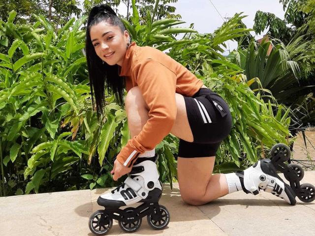 TrinityPrestige, crouched on a concrete path, wearing an orange hoodie, black shorts, and white rollerblades with black accents. She has long black hair and a flirtatious smile. Lush green plants in the background.