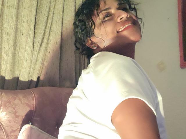 Fotografía de IvannaDuran con cabello rizado oscuro, sonriendo a la cámara, vistiendo una camiseta blanca. La luz del sol ilumina su rostro y parte superior del cuerpo. Su cabeza está ligeramente inclinada, creando una atmósfera juguetona..