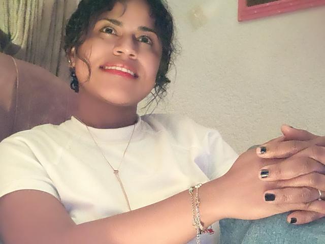 Fotografía de IvannaDuran con cabello oscuro y rizado, sonriendo, vistiendo una camiseta blanca, esmalte de uñas negro y un pulsera, manos descansando sobre un cojín azul, sutil tensión sexual.