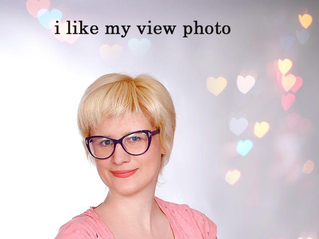 Fotografia de SabrinaShine, uma mulher loira com cabelo curto, usando óculos de aro preto e uma camisa rosa, sorrindo. Contra um fundo branco, com luzes bokeh coloridas. O texto acima diz "Eu gosto da minha foto da vista".".