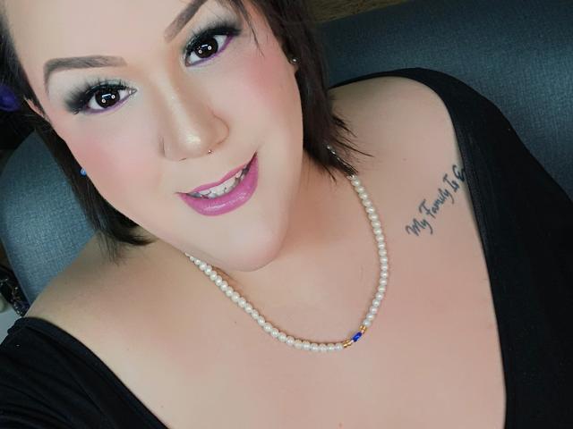 Close-up van GiiannaMorrys, een mollige vrouw met lichte huid, zwart haar en veel make-up, die een zwarte top draagt met een open schouder. en een witte parelketting. "Mijn hart klopt" tatoeage op haar borst. Verdomd sexy glimlach..