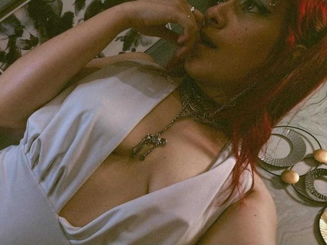 Fotografía de primer plano de LolyYunno con cabello rojo, mordiéndose el dedo, vistiendo un vestido blanco de escote pronunciado que revela un amplio escote. collar de plata con forma de cruz, y collares en capas, que transmiten una intensa tensión sexual..