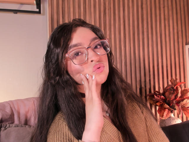 Eva McAllister, una mujer atractiva con cabello negro largo y gafas de montura clara, hace un puchero con la mano en la mejilla. Ella lleva una blusa de punto marrón, el fondo tiene listones de madera y plantas rojas..