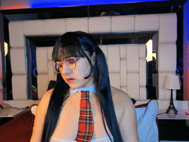 SaraFountain, with long black hair and glasses, wears a white strapless top and red plaid necktie, looking down, in a dimly lit room with a tufted headboard and lamps. Fucking sexy.