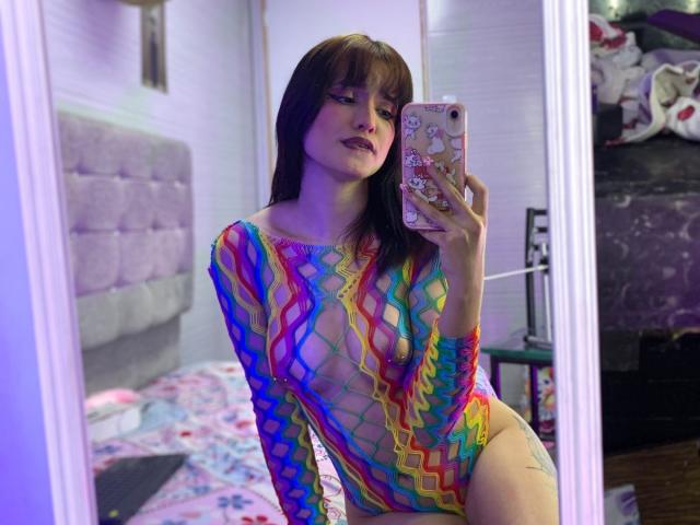 Mitsury Kanrojy, avec sa peau pâle, ses cheveux noirs et sa frange, portant une combinaison transparente et colorée en résille, tenant un téléphone dans sa chambre avec des lumières vives, mettant en valeur un grand pénis. Miroir réfléchissant.