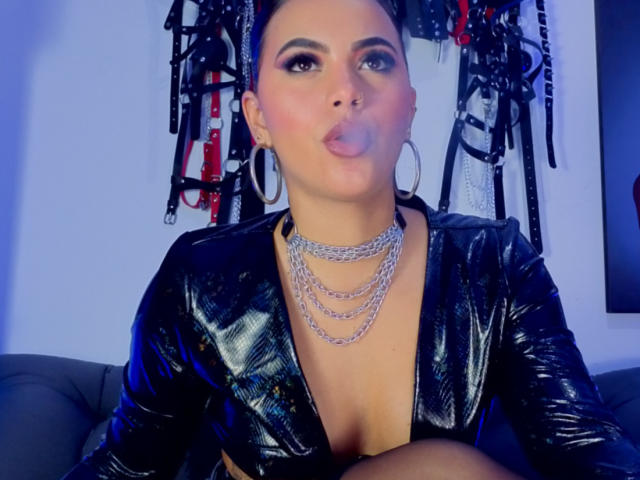 KateStones tem uma pele clara, cabelo escuro em tranças, veste uma blusa de couro preto brilhante, um colar e está soprando. Uma bolha de chiclete. Equipamentos de BDSM estão pendurados ao fundo..