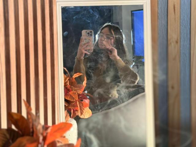 El reflejo en un espejo muestra a EvaMcAllister tomando una selfie con un teléfono, vistiendo una sudadera marrón, rodeada de hojas de otoño. hojas, con vapor en el espejo, luz azul en el fondo.