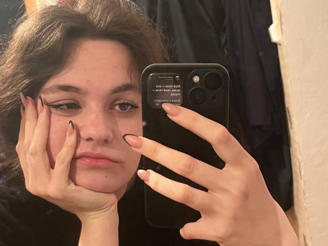 LahomaLoven with fair skin, dark wavy hair, and black eyeliner takes a selfie with a black iPhone, holding her face with both hands. Her expression is neutral, with visible moles. Fucking sexy.
