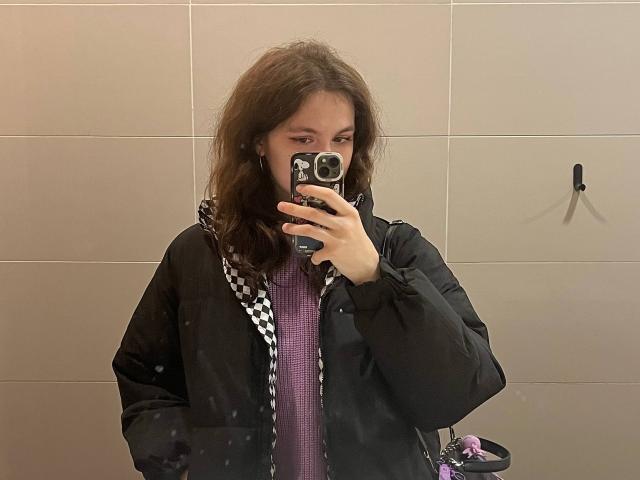 LahomaLoven with wavy brown hair takes a mirror selfie, holding a black phone with a black case, wearing a black jacket over a purple checkered shirt, in a beige-tiled room.