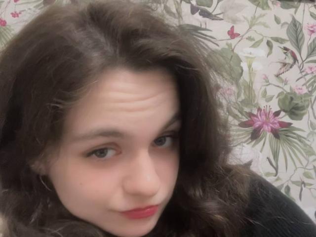 Close-up of LahomaLoven with wavy brown hair, pale skin, and red lips, gazing directly at the camera. Floral pattern in background. Fucking intense expression, slightly tilted head.