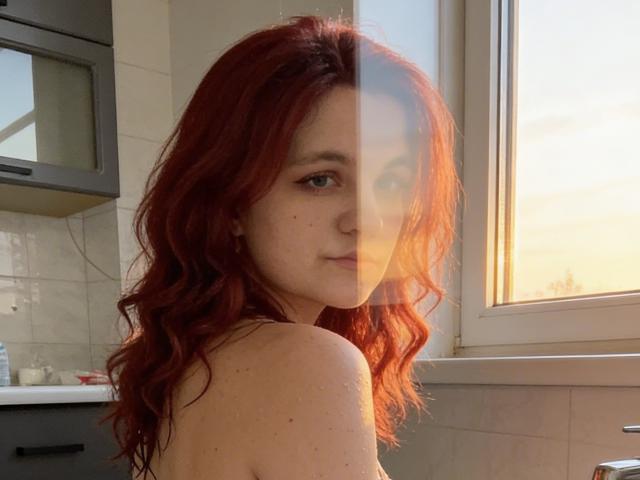 Fotografía de LahomaLoven con cabello rojo largo y ondulado, mirando por encima del hombro, desnuda, con pecas claras, luz natural suave. Desde la ventana, se ve un fondo con armarios de cocina tipo azulejos, con un ambiente casual, sexy y seductor..