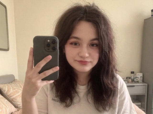 LahomaLoven, con i capelli castano scuro spettinati, tiene in mano un iPhone nero in un selfie, indossando una camicia bianca. Lei sorride leggermente, con un trucco leggero, in una camera da letto con un letto e un comodino. Molto attraente..