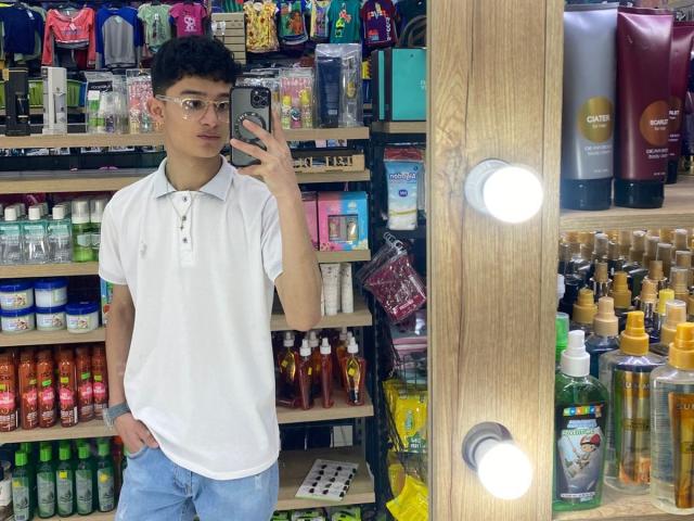 Patrick Holland está de pie en una licorería, vistiendo una camisa polo blanca y pantalones vaqueros azules, sosteniendo un teléfono, con la mano en el bolsillo. rodeado de estanterías llenas de botellas de diferentes tipos, con luces brillantes que resaltan la escena..