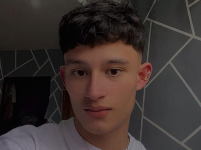 Selfie em close-up de PatrickHolland, um jovem com cabelo preto curto, pele clara e olhos castanhos. Usando uma camiseta branca, com um brinco na orelha esquerda. O fundo tem um padrão geométrico de azulejos cinzas. Rosto incrivelmente atraente..