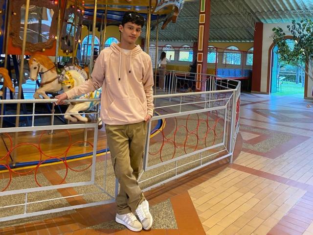 Patrick Holland, een jonge man met lichte huid, een zwarte pet, een roze hoodie en beige broek, leunt tegen een metalen railing in de buurt van een een carrousel, zijn penis is niet zichtbaar, staand in een helder verlichte, overdekte ruimte..