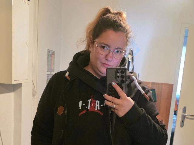 Fotografie von Sarah-kommt-hot mit einem Pferdeschwanz, die eine Brille trägt und eine schwarze Kapuzenjacke mit dem Aufdruck "R." trägt. Eine Person, vermutlich eine Frau ("I. P."), hält ein Smartphone, steht in einem hell erleuchteten Raum und schaut in die Kamera..