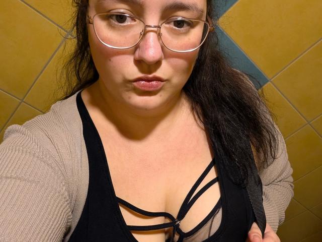 Fotografía de primer plano de Sarah-kommt-hot, una mujer latina con gafas, un sujetador negro con detalles de tiras y un cárdigan beige. mirando directamente a la cámara, fondo con baldosas amarillas, expresión intensa, ligera abertura en el escote..