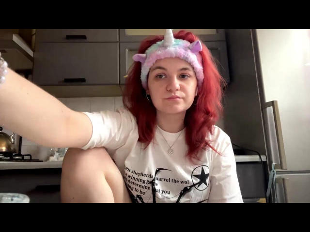 LahomaLoven, met rood haar, draagt een roze eenhoornhoofdband en een wit shirt met zwarte tekst, zit in een keuken en strekt zich uit. Een hand richting de camera. Zichtbaar dijbeen, subtiele seksuele spanning.
