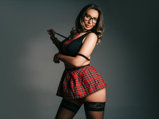 GinaBear se tient face à la caméra, tenant une sangle noire contre son épaule. Elle porte une mini-jupe rouge à carreaux, un haut en dentelle noire et des bas résille montants jusqu'aux cuisses. Elle porte des lunettes, avec une expression séduisante..