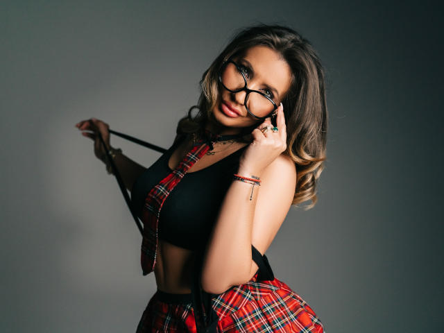 GinaBear mit schulterlangem, gewellten Haar, trägt ein schwarzes Crop-Top und einen roten karierten Rock, hält einen schwarzen Träger und justiert ihn. Brille, verführerischer Blick, dunkles, schattiges Gesicht, sexy Pose, verführerischer Ausdruck..