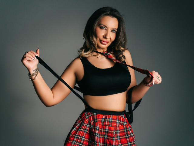 GinaBear, com pele morena média e cabelo castanho escuro, vestindo uma cropped preta e uma saia xadrez vermelha, segurando um objeto preto suspensórios. Ela tem um olhar sedutor, que enfatiza seus grandes seios..