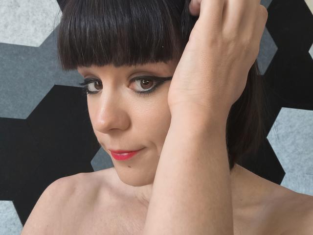 Close-up van OrianaLaFrancaise met een kort zwart kapsel, rode lippenstift en donkere eyeliner. Ze heft haar rechterhand en raakt haar hoofd aan. Zwarte en witte geometrische achtergrond. Heel aantrekkelijk..