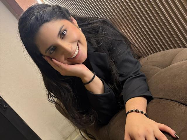 Luciana Blake, met lang zwart haar, leunt tegen een bruine bank en glimlacht, met haar hand op haar wang. Ze draagt een zwart shirt, zwarte nagellak en een zwarte armband. Heel erg aantrekkelijk.