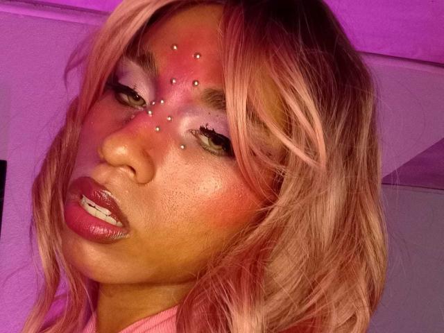 Nahaufnahme eines Fotos von VenusTsHot mit rosa Haaren, viel Make-up und roten, leuchtenden Gesichtspiercings. Sie hat volle, glänzende Lippen und einen verführerischen, leicht geöffneten Blick. Ein neonfarbenes, violettes Licht wirft einen verführerischen Schein..