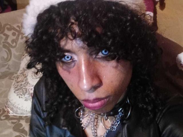 Fotografia ravvicinata di VenusTsHot, una donna nera con capelli ricci e occhi azzurri, che indossa una giacca nera e un collare con borchie. con catene. Lei alza lo sguardo con un'espressione seria.