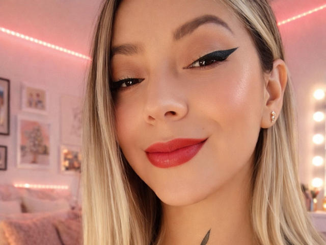 Fotografía de primer plano de KayleeM con piel clara, cabello rubio, delineador de ojos negro alado y lápiz labial rojo. Ella está sonriendo sutilmente, con una sonrisa arrogante. Iluminación suave, fondo borroso de un dormitorio con cuadros enmarcados..