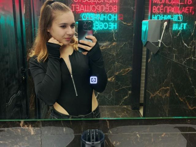 DenniseDaghita, brunette with ponytail, wearing black zip-up top, taking mirror selfie in dark bathroom with neon signs. She has a sexy, confident expression, phone in hand, black marble tiles.