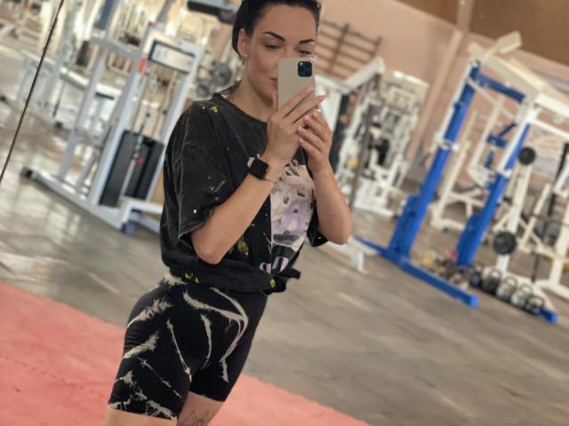 Xioni staat in een sportschool en maakt een selfie met een smartphone. Ze draagt een zwart shirt en zwarte korte broek met witte patronen, en kijkt iets opzij, met een sexy, zelfverzekerde uitstraling. sfeer.