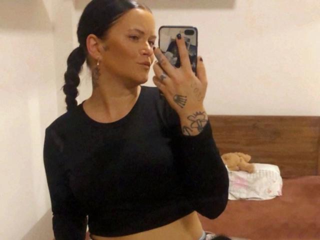 NicolSparkleX prend un selfie devant un miroir, tenant un téléphone et montrant ses mains tatouées. Elle porte un crop top noir, et ses cheveux sont tressés. Un ours en peluche est sur un lit en arrière-plan..