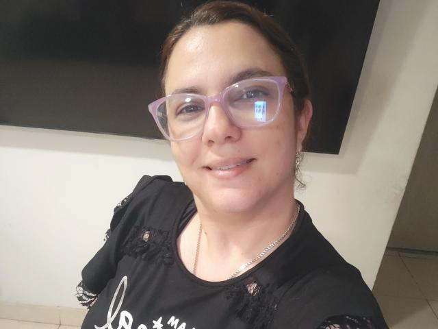 Amelia Wilson, com cabelo castanho, usando óculos de grau e uma blusa preta com detalhes em renda, sorrindo levemente. Ela está olhando diretamente para a câmera, usando um colar de prata. Iluminação brilhante.