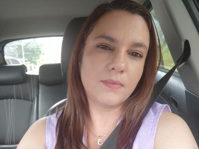 Close-up de Amelia Wilson dentro de um carro, suando, com cabelo castanho, camiseta branca e cinto de segurança. Ela tem uma expressão neutra, com luz natural vinda da janela. Focada em seu rosto.