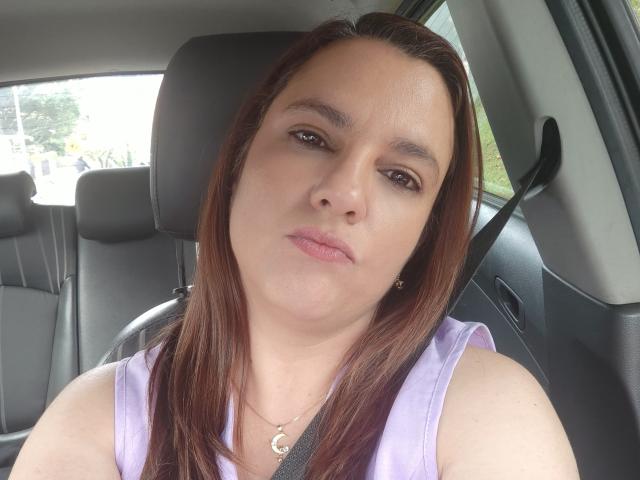 Close-up of AmeliaWilson in a car, seated with seatbelt across chest. She has long brown hair, light skin, wearing a white tank top, gazing directly at the camera with a neutral expression.