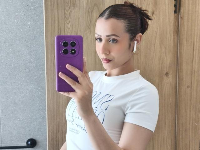AliaahSky tira uma selfie no espelho, segurando um telefone roxo com três câmeras. Ela está vestindo uma camisa branca, tem o cabelo escuro preso em um coque e está usando fones de ouvido brancos. Muito quente.
