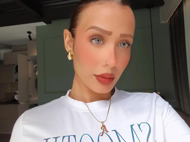 Close-up photo of AliaahSky, a fair-skinned woman with blue eyes, red lipstick, and a nose ring. She's wearing a white shirt, gold earrings, and a gold necklace. Bright light highlights her face.