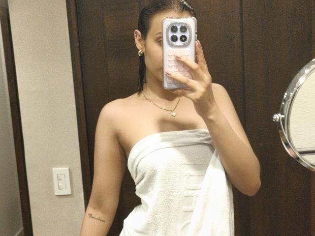 AliaahSky, con el cabello oscuro y mojado, está de pie en un baño, tomando una foto de sí misma con un iPhone usando un espejo. Está envuelta en una toalla blanca, cubriendo sus senos, y tiene un tatuaje en su brazo derecho..