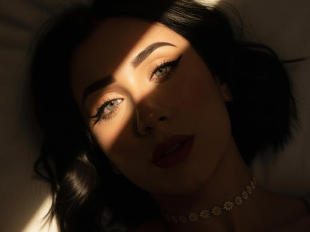 Gros plan d'AliaahSky avec des cheveux foncés, éclairée par la lumière du soleil, créant des ombres saisissantes sur son visage. Elle porte un collier de fleurs, avec un maquillage discret, et une expression séduisante. Une lumière douce met en valeur ses yeux..