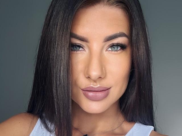 Gros plan de LizaDream avec des cheveux noirs lisses, des yeux verts et des lèvres roses pulpeuses. Elle porte un haut blanc et regarde directement l'appareil photo avec un sourire subtil et séduisant. Fond gris.